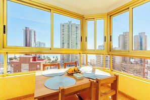 Una stanza d'angolo con un'ampia finestra, un tavolo in legno con una ciotola di frutta e vista su edifici alti.