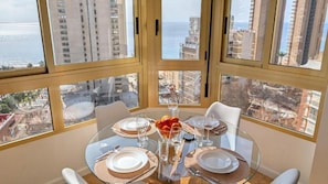A dining area with a glass table, white chairs, and a view of the ocean and buildings. - Gerona Apartments (Benidorm)