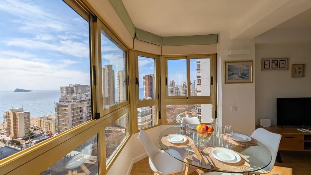 Um apartamento alto com varanda, janelas amplas, área de jantar com mesa de vidro e uma televisão de tela plana.