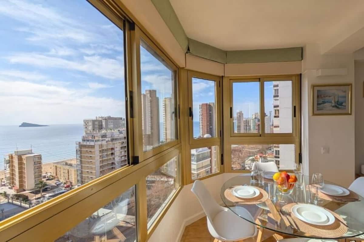 Um apartamento alto com varanda, janelas amplas, área de jantar com mesa de vidro e uma televisão de tela plana.