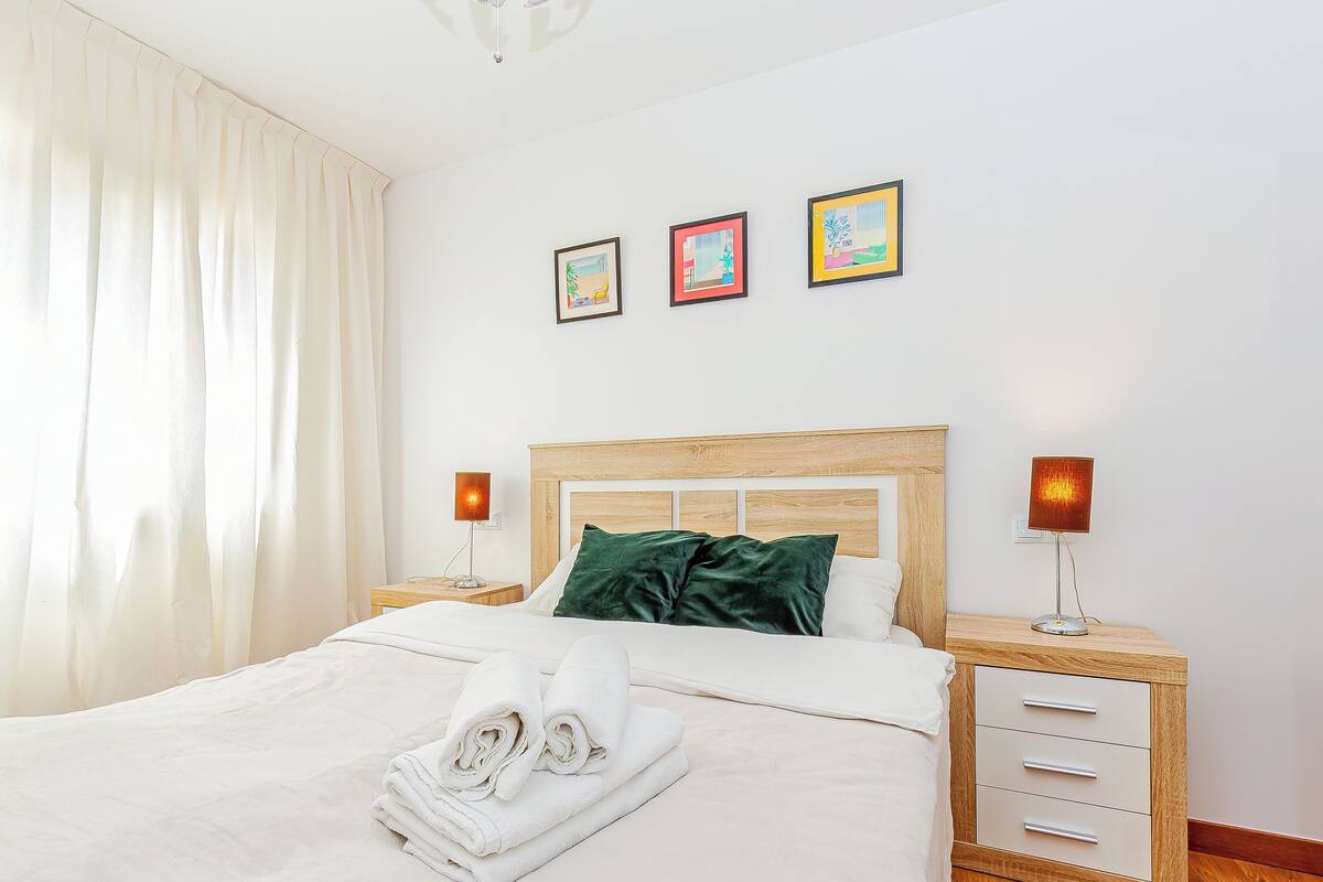 A neatly made bed with pillows, a bedside lamp, and a nightstand with a lamp. There are three framed pictures on the wall.