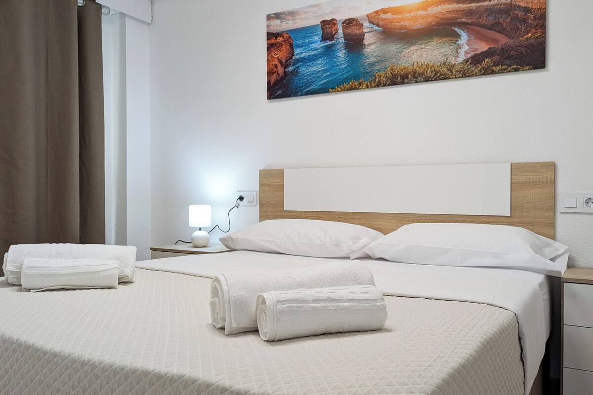 A neatly made bed with white linens and towels, a bedside lamp, and a painting of a coastal landscape on the wall.