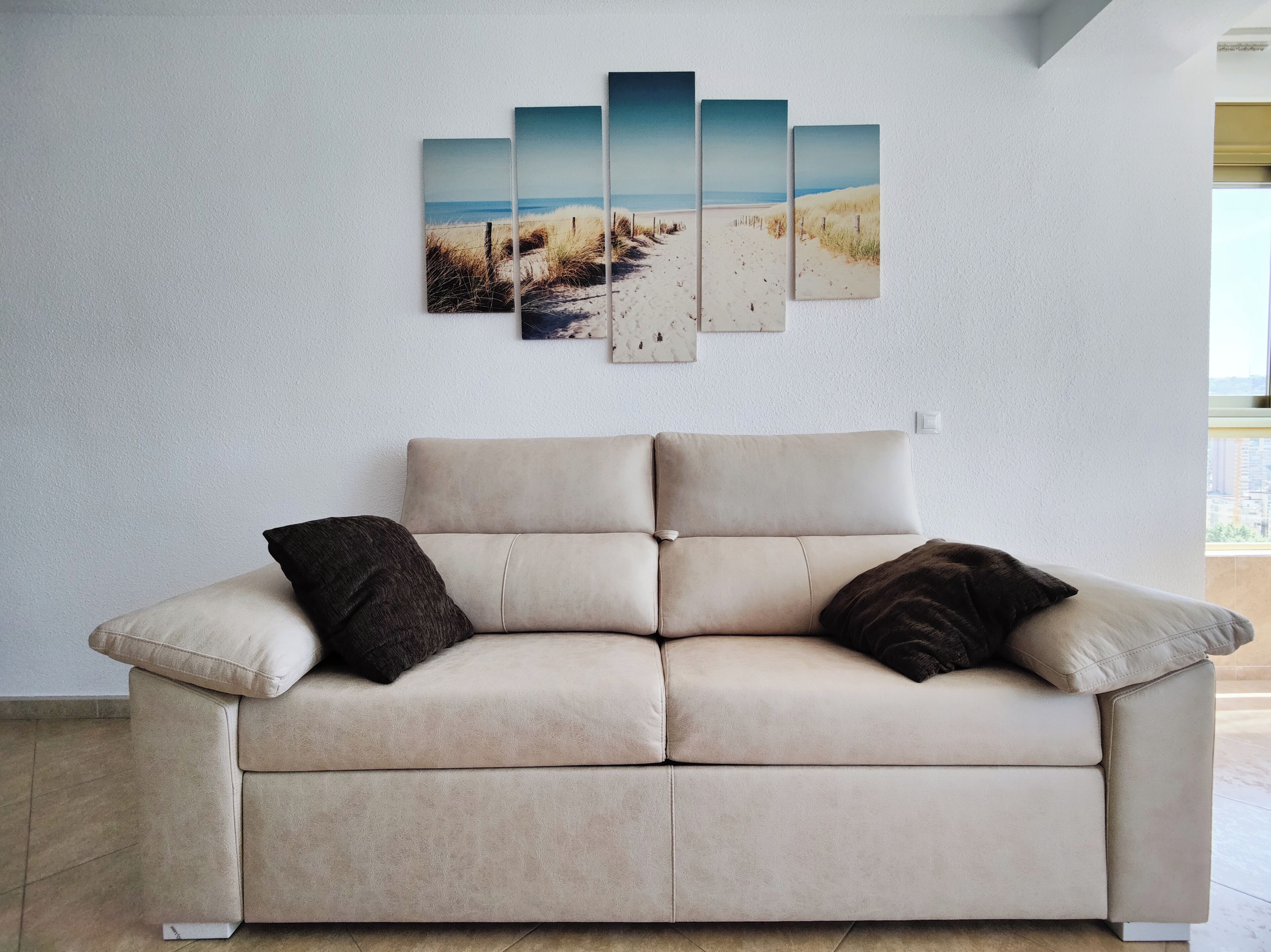 A beige sofa with two dark cushions, a white wall, and a beach scene artwork.