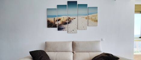 A beige sofa with two dark cushions, a white wall, and a beach scene artwork.