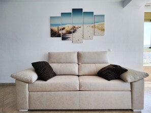Un canapé beige avec deux coussins foncés, un mur blanc et une œuvre d’art représentant une scène de plage.