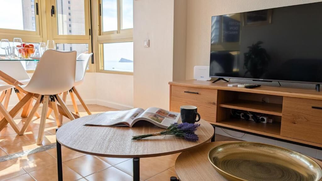 Una sala moderna con un televisor de pantalla plana grande, una mesa de comedor con sillas, una mesa de centro con un libro y flores, y un mueble de madera que funciona como base para el televisor.