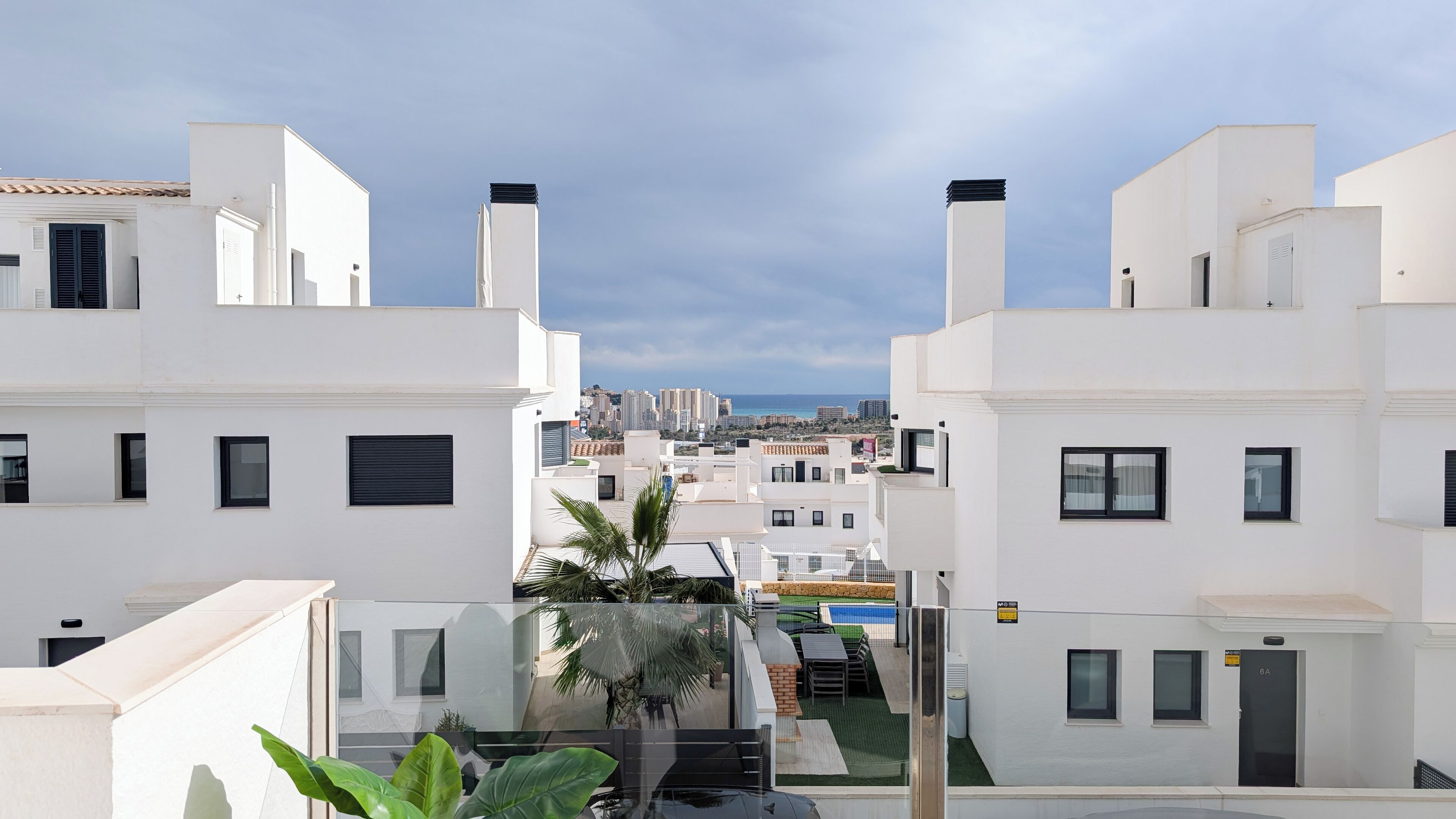 A modern residential area with white buildings, a visible pool, and a clear view of the sea.