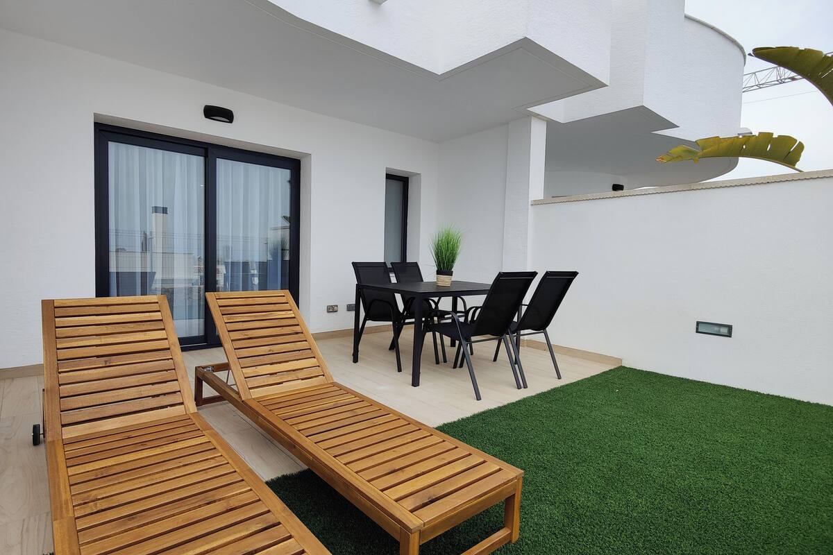A patio with two wooden lounge chairs, a dining table with chairs, and a small garden area with artificial grass.