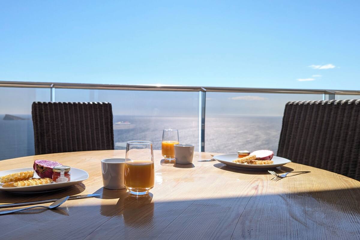 Uma mesa com pratos de comida, copos de suco e uma garrafa de água em uma varanda com vista para o oceano.
