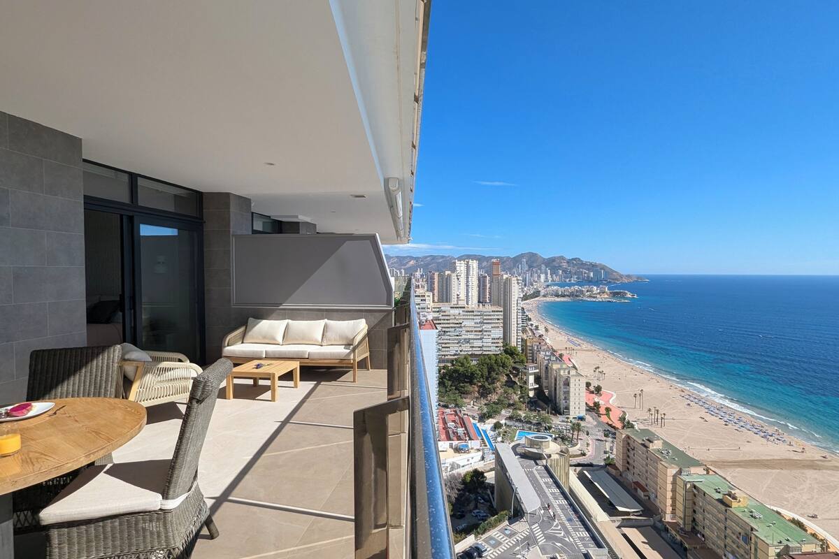 Varanda com mobiliário de vime, uma mesa com prato e copos, e vista para a praia e o horizonte da cidade.