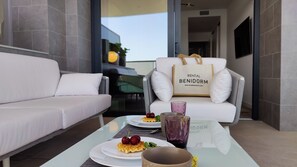 A modern interior with a white sofa, a glass table with plates of food, a chair with a 'RENTAL BENIDORM' bag, and a TV mounted on the wall. - Sunset Cliffs Apartments Tower 1 (Benidorm)