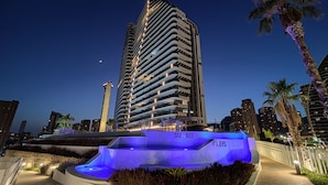 A high-rise building with a modern design, a well-lit entrance, and a landscaped area with a water feature. - Sunset Cliffs Apartments Tower 1 (Benidorm)