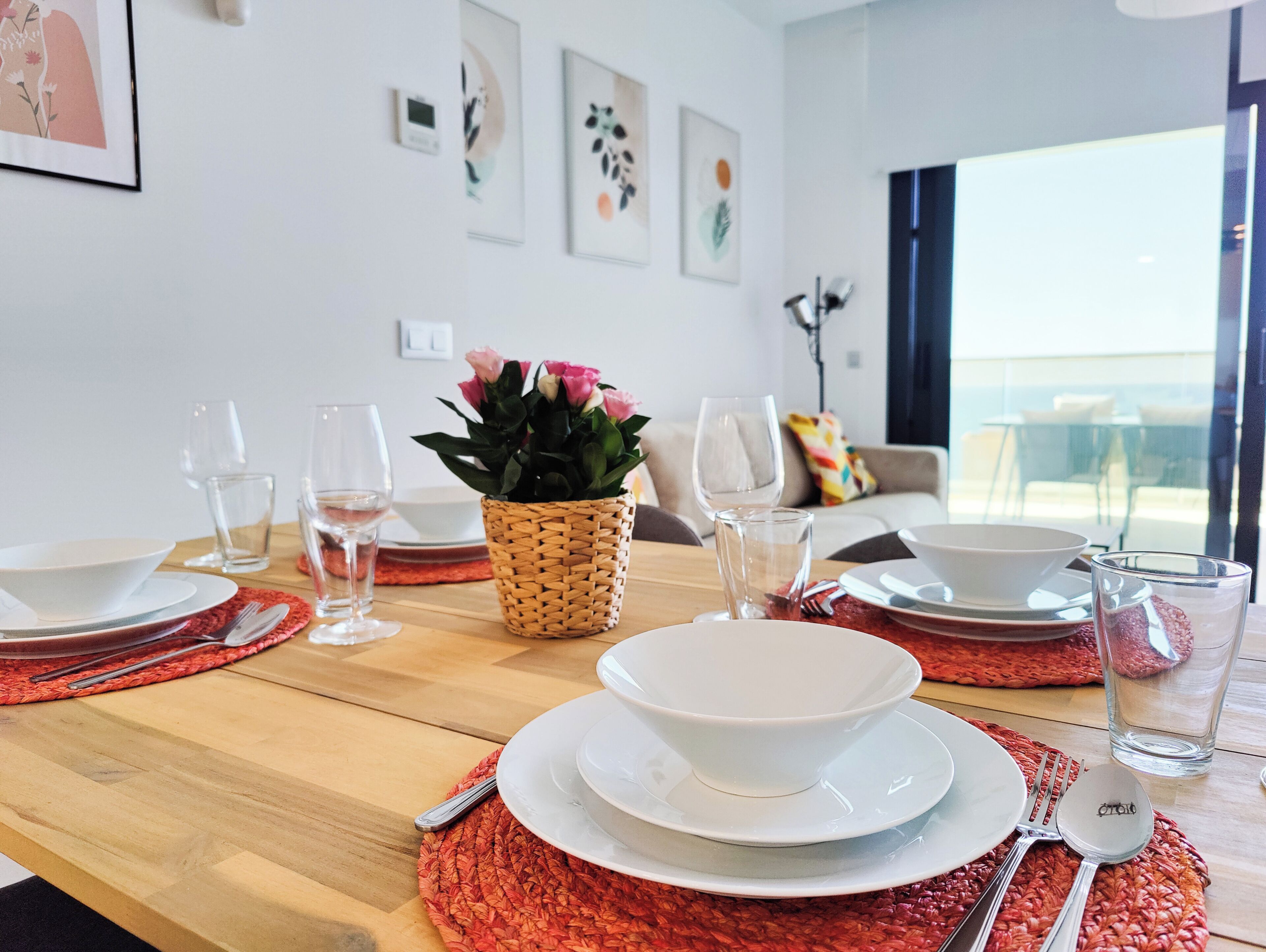 Une table à manger dressée avec des assiettes, des bols, des couverts et des verres, agrémentée d’un set de table en fibres tressées et d’une plante en pot.
