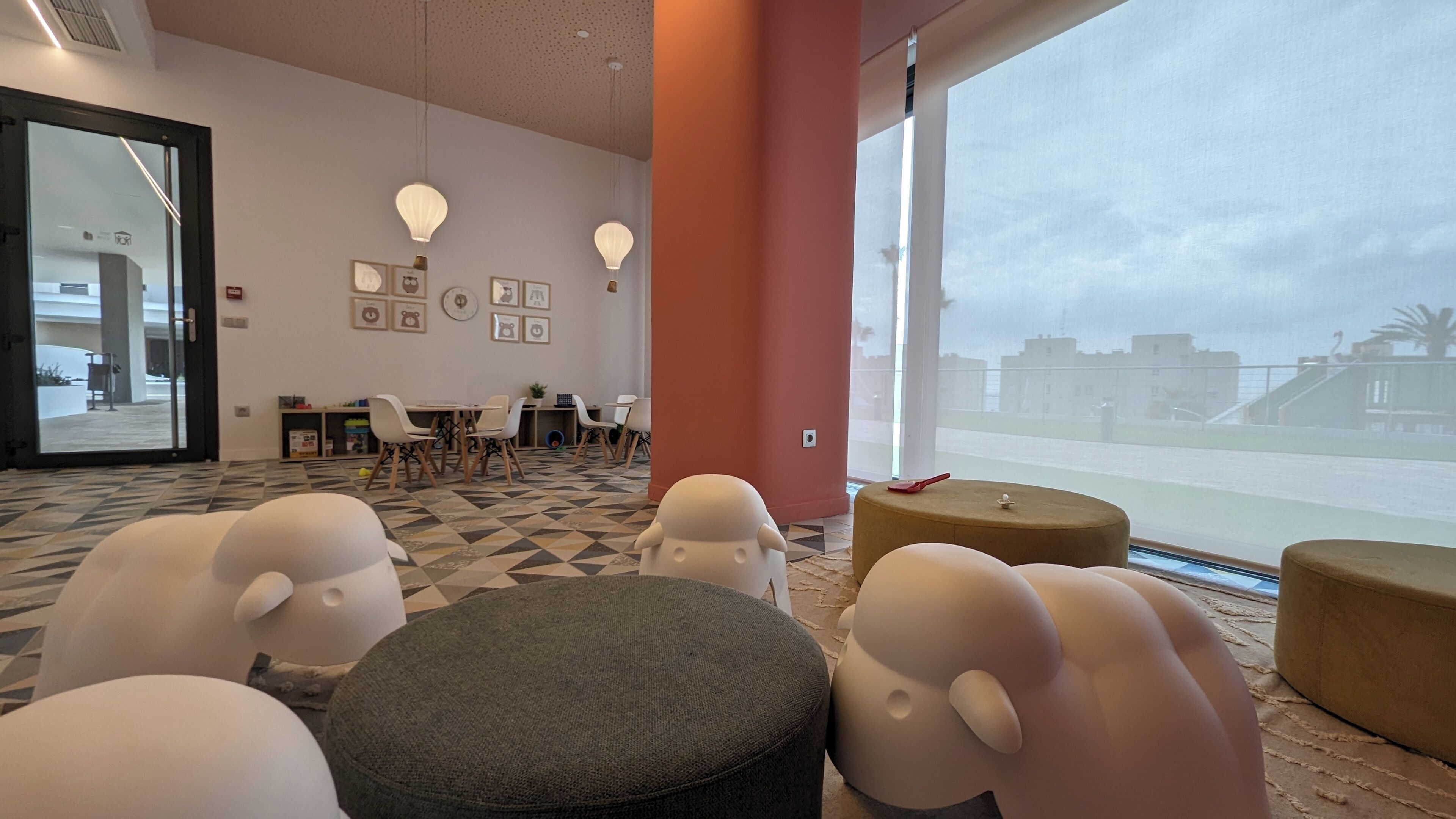 Un vestíbulo de hotel con un gran ventanal, una zona de reuniones con mesas y sillas, y un área de juegos para niños con asientos cómodos.