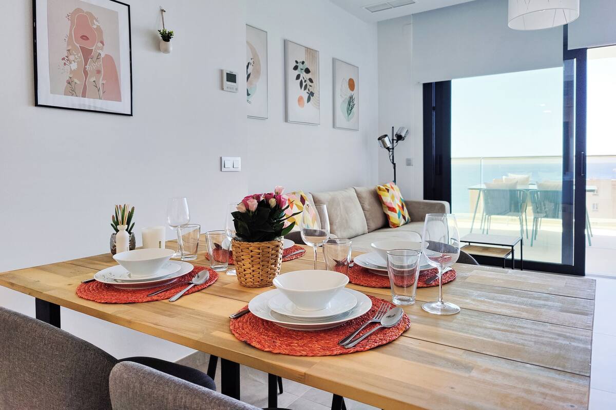 Uma área de refeições com uma mesa de madeira posta para quatro pessoas, um sofá e vista para o mar.