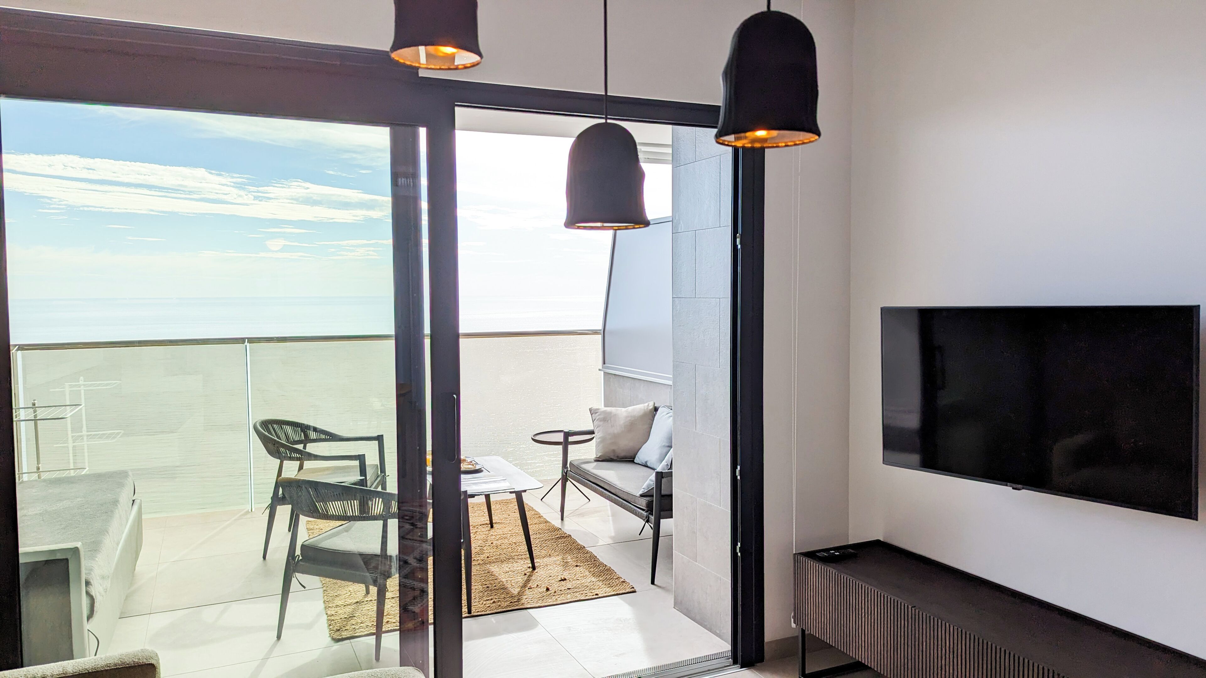 A modern balcony with a glass door, a sofa, a small table, and a TV mounted on the wall.