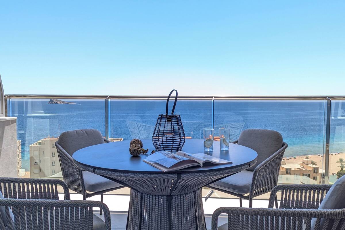 Ett runt bord med flätade stolar, en svart vävd väska och ett glas vatten på en balkong med utsikt över havet.