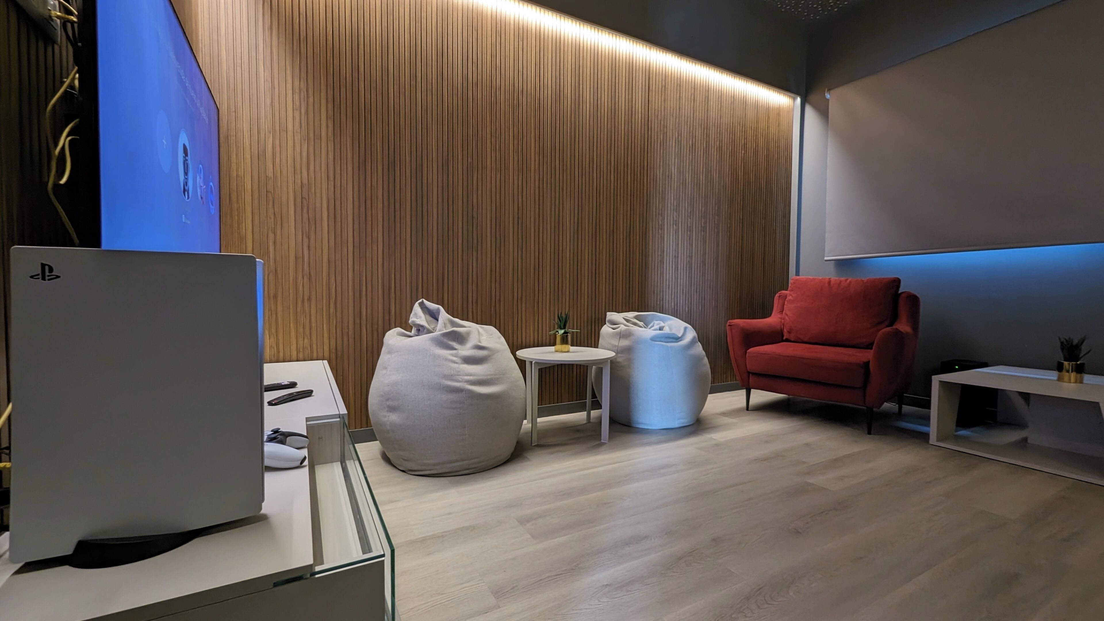 A modern room with a wooden slatted wall, a red chair, a white bean bag, a small round table with a plant, and a white desk with a computer.
