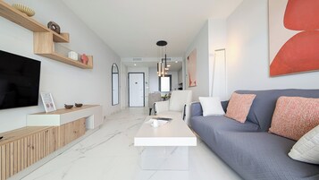 Uma sala de estar moderna com uma TV de tela plana, uma mesa de centro, um sofá com almofadas e uma área de jantar ao fundo.