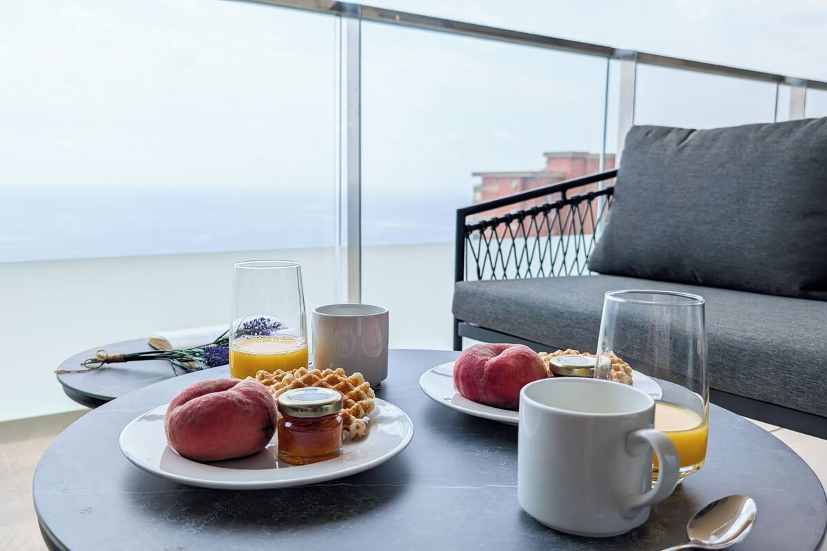 En balkon med et bord dækket til morgenmad, inklusive kager, et glas appelsinjuice og et lys.