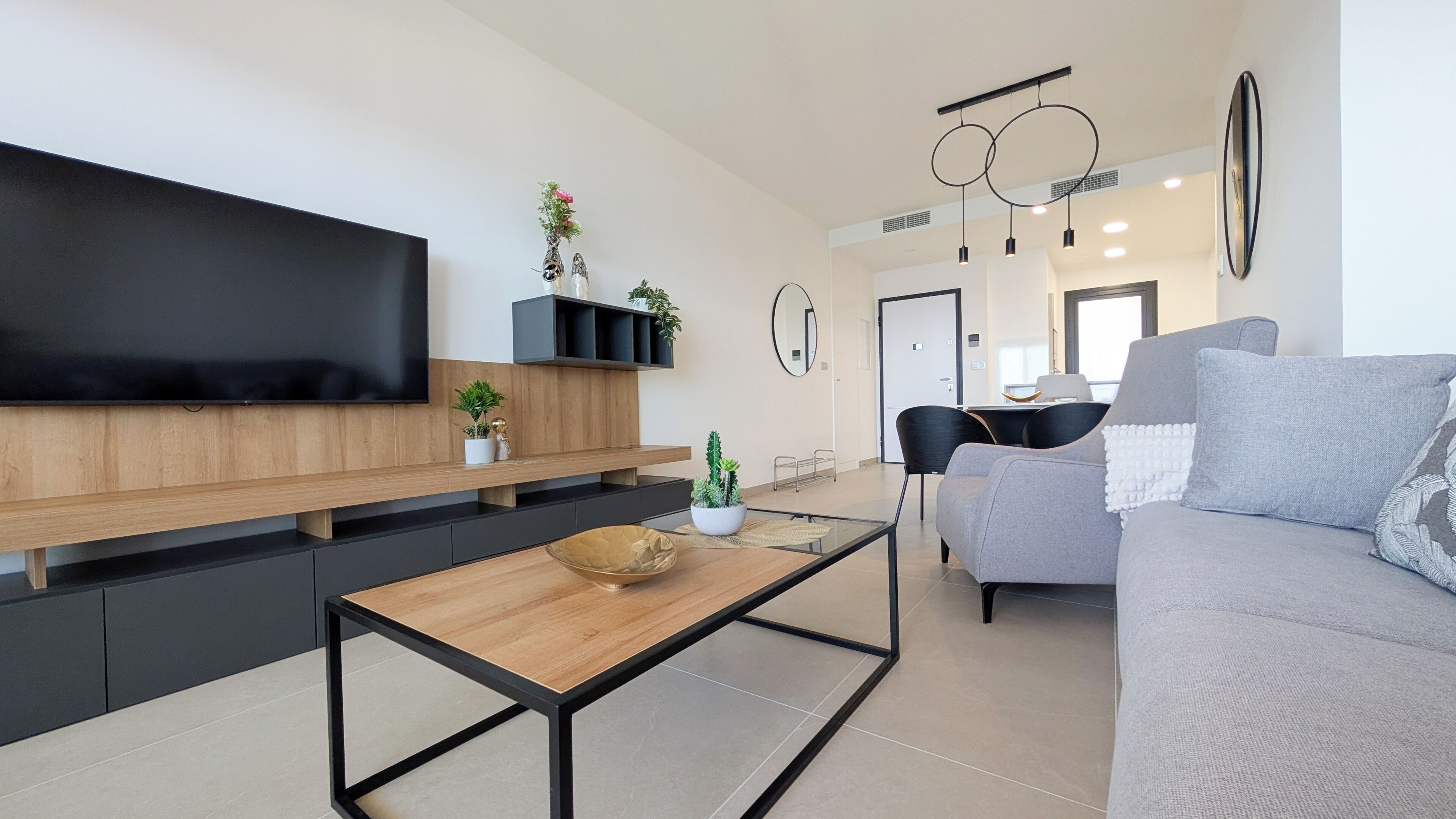 Un salon moderne avec un téléviseur à écran plat, une table basse en bois, un canapé gris et un coin repas à l’arrière-plan.