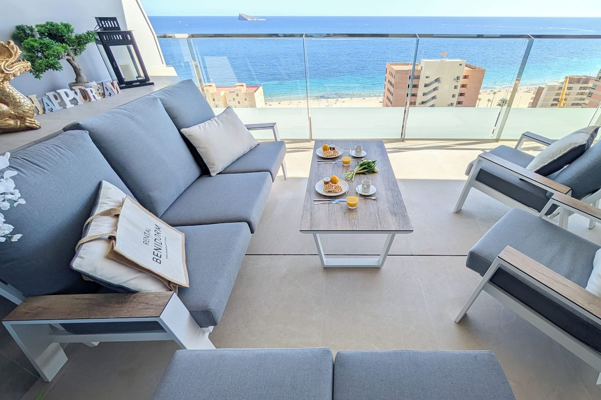 En balkon med sofa, sofabord og udsigt til strand og hav.
