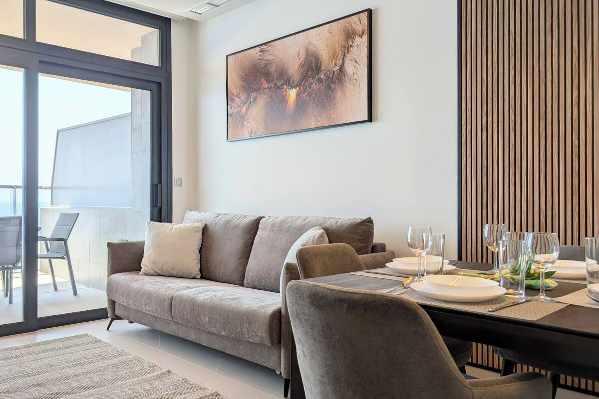 En moderne stue med sofa, spisebord til to og et stort vindue med udsigt til en balkon.