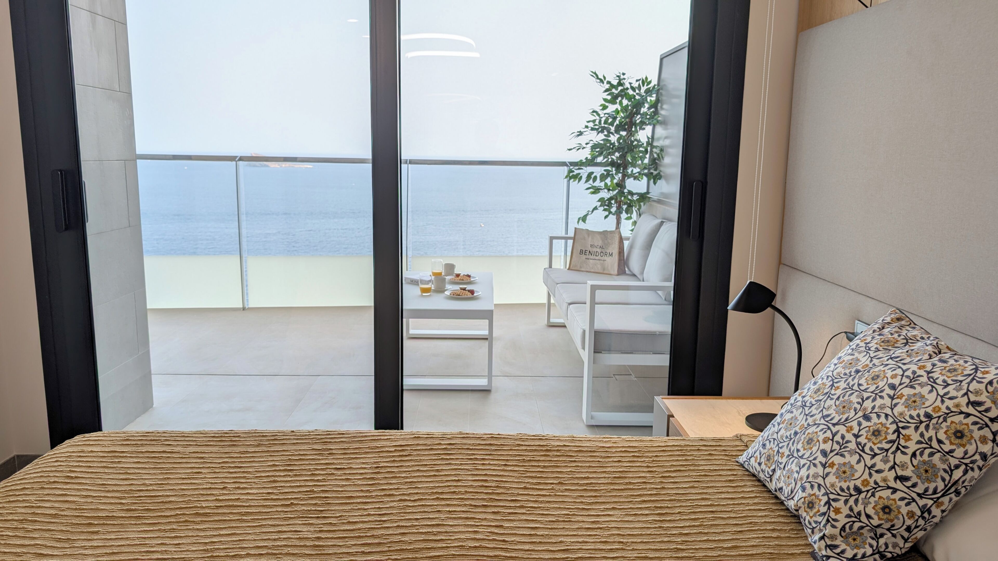 Habitación de hotel con cama, escritorio, silla, una mesita con artículos de desayuno, una planta y vista al mar.