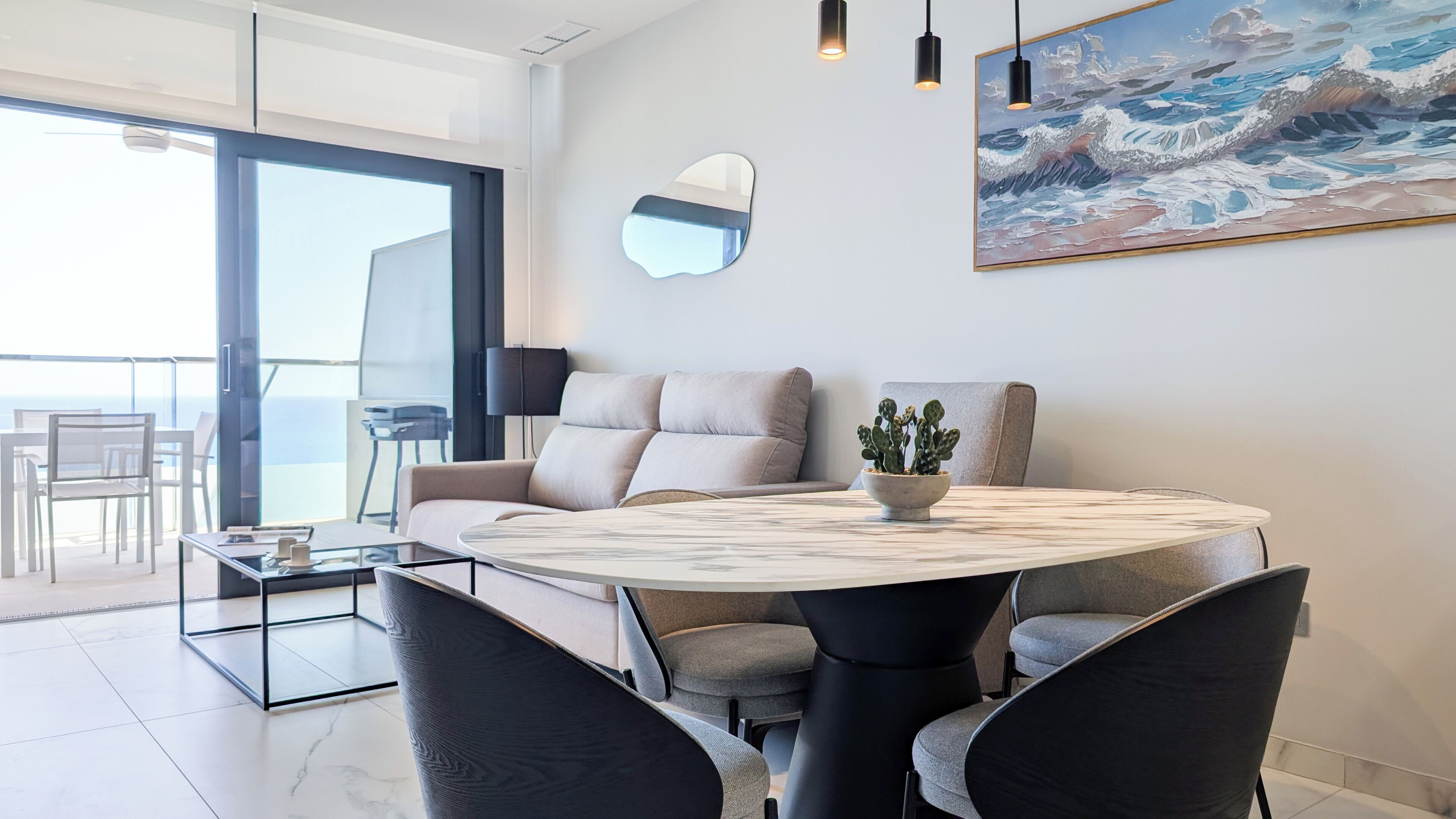 A modern living room with a round table, grey sofas, a cactus plant, a painting, and a large window overlooking a balcony.