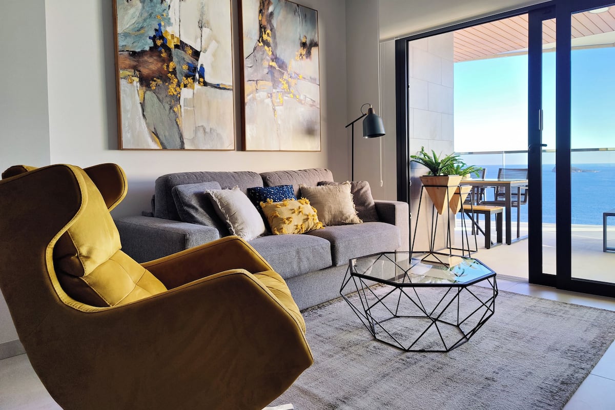 En moderne stue med sofa, sofabord og havudsigt gennem store glasdøre.