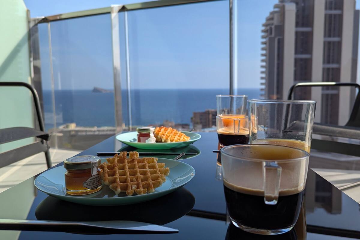 Et bord med vafler, et kaffeglas og en honningflaske på en balkon med udsigt over havet og bygninger.