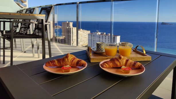 A balcony with two plates of croissants and orange juice, a wooden tray, and a view of the ocean and cityscape. - Sunset Waves Apartments Tower 1 (Benidorm)
