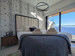 Une chambre moderne avec un grand lit, un fauteuil suspendu près de la fenêtre et une vue sur la mer.