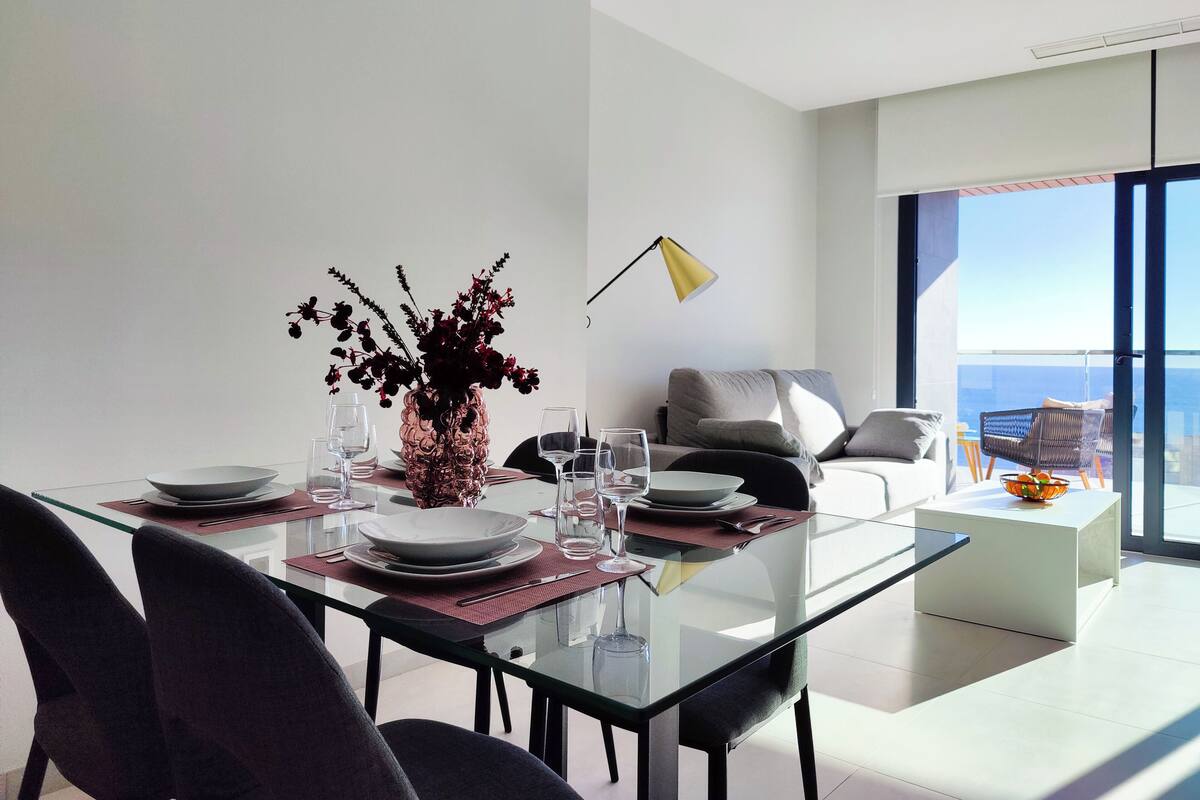 A modern dining area with a glass table, dark chairs, and a vase of flowers. There is a sofa and a chair in the background with a view of the sky.