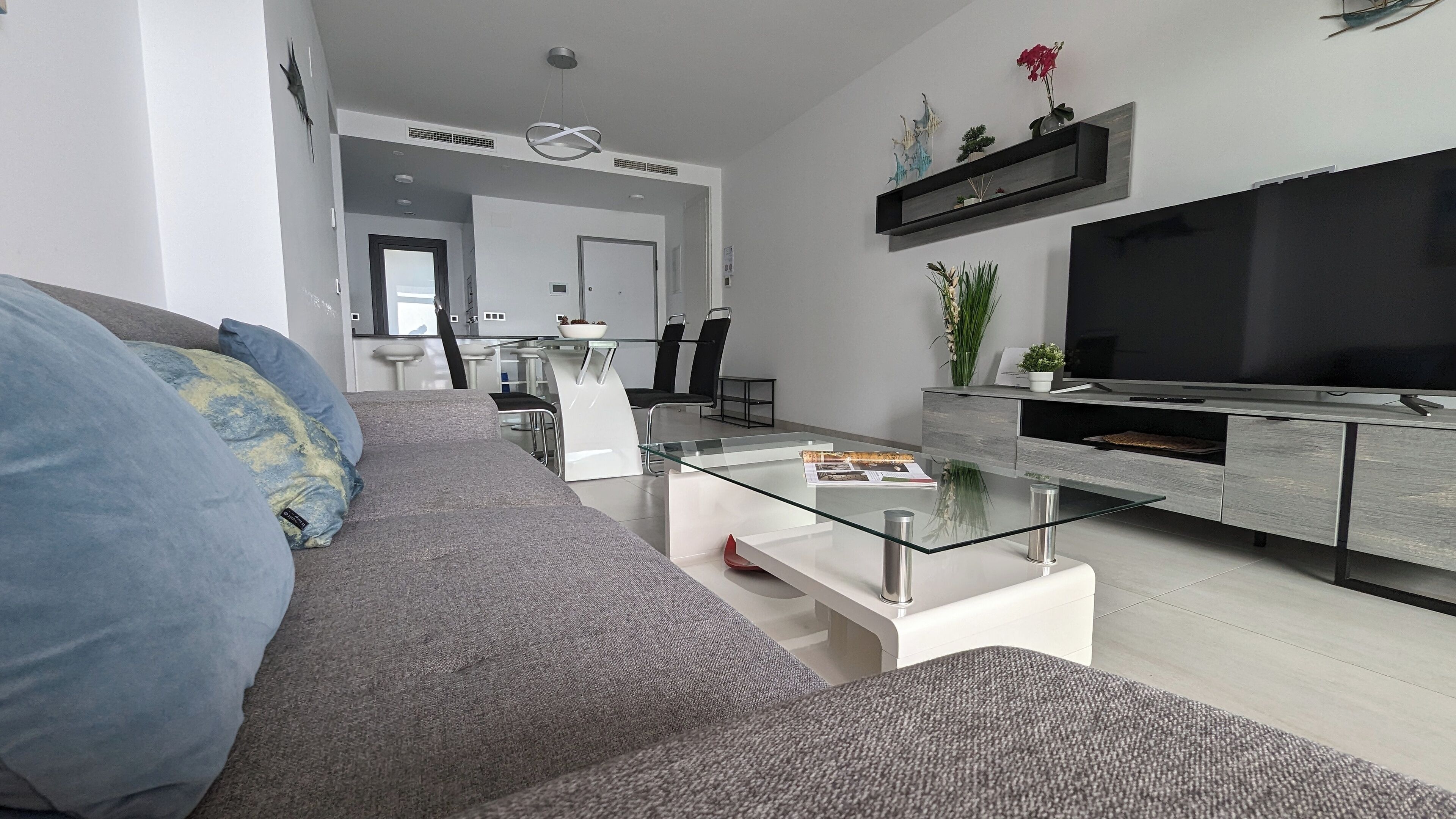 Una sala de estar moderna con un sofá gris, una mesa de centro de vidrio y un televisor sobre un mueble de madera.