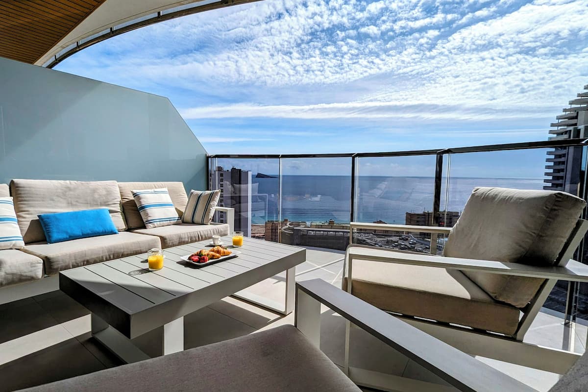 A modern outdoor seating area with a sofa, chairs, and a table, overlooking a coastal cityscape.