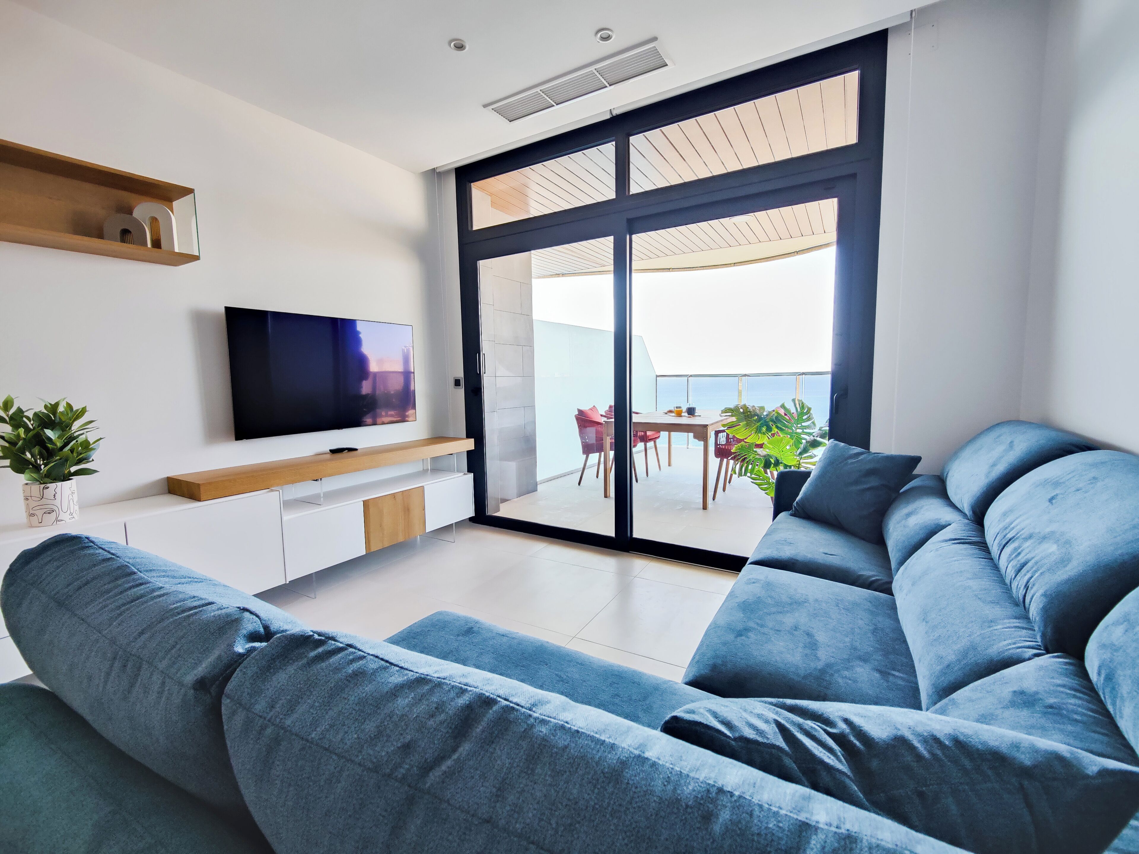 A modern living room with a large blue sofa, a flat-screen TV on a wooden console, and a sliding glass door leading to an outdoor dining area with red chairs.