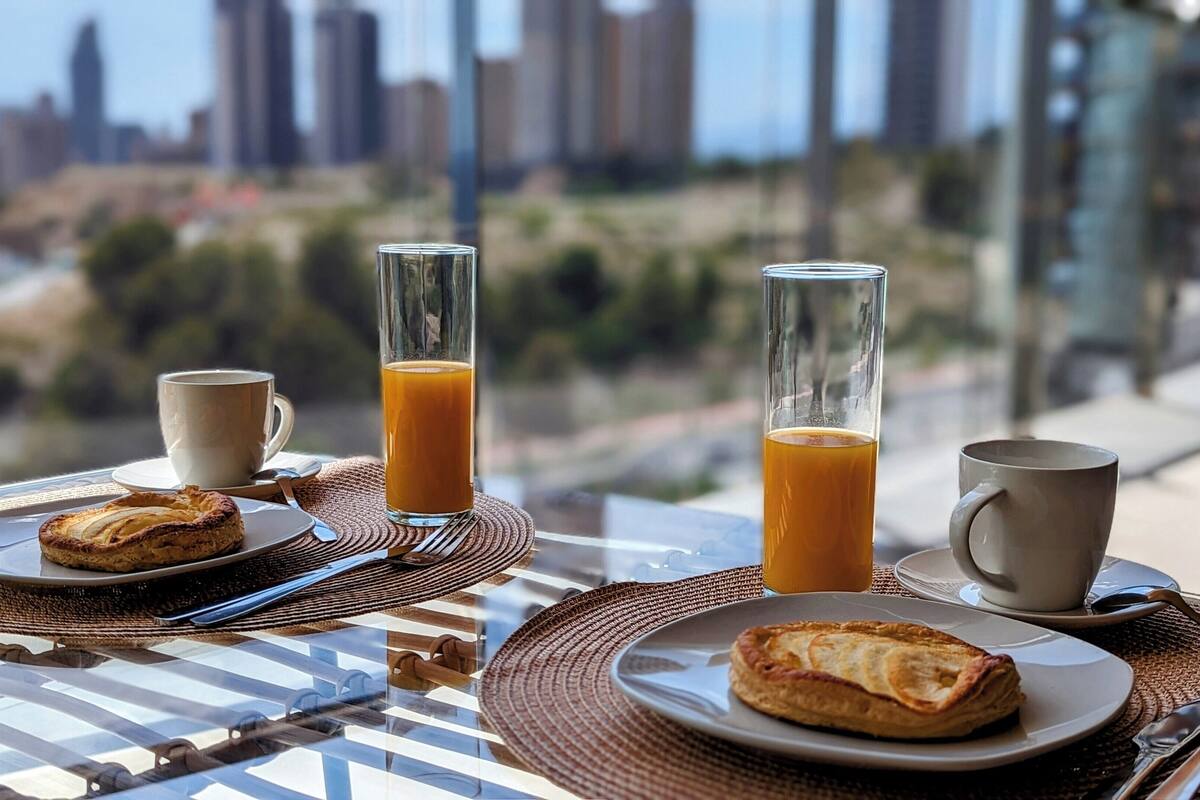 A breakfast setting with two pastries, two cups of coffee, and two glasses of orange juice on a table with a city view.