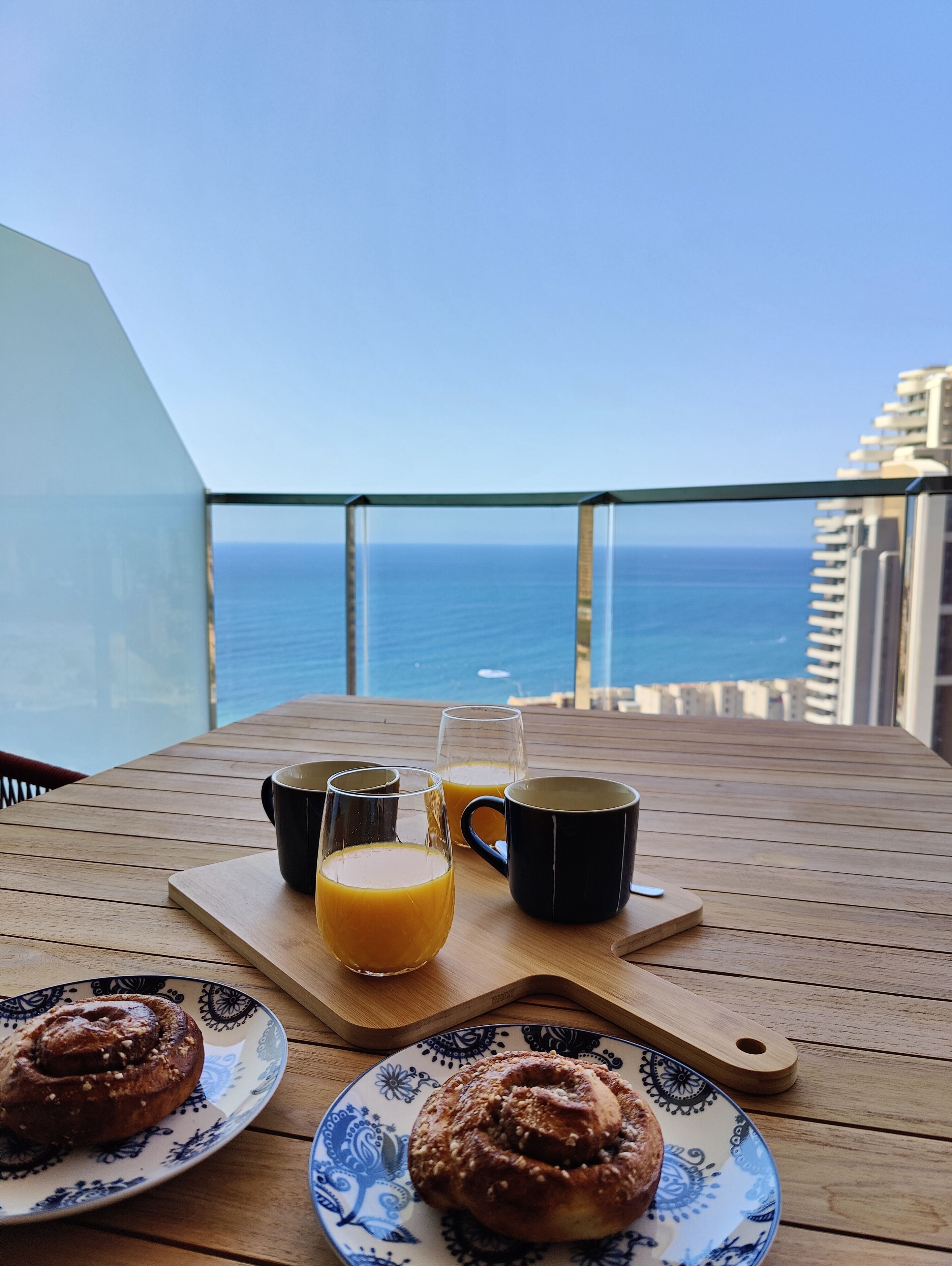 Une table en bois, dressée sur un balcon donnant sur l’océan, est garnie de divers éléments pour le petit-déjeuner, notamment deux viennoiseries, deux tasses et deux verres de jus d’orange.