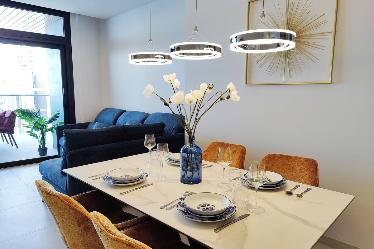 A dining area with a white table set for four, surrounded by chairs, a vase with flowers, and modern pendant lights.