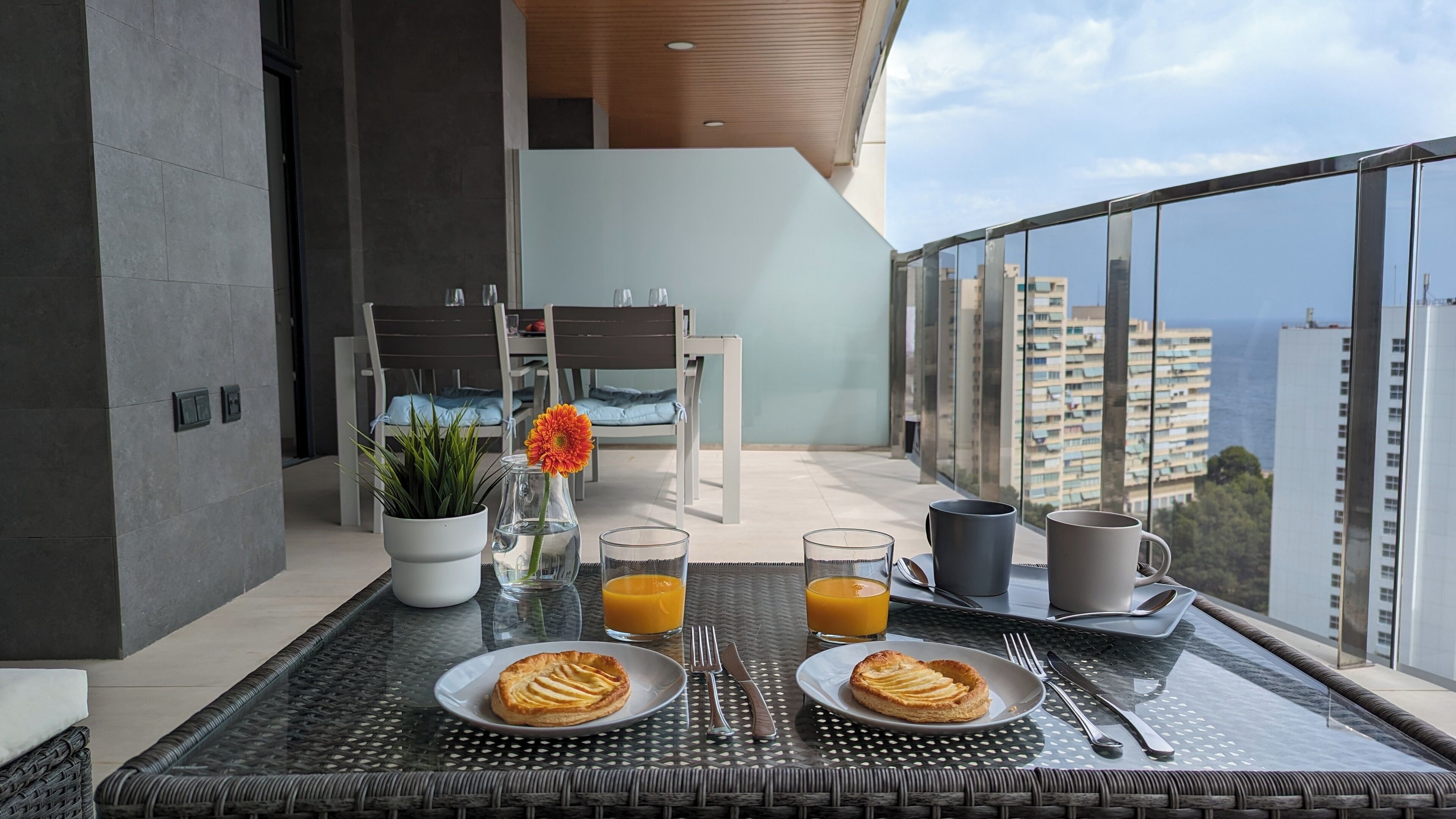 Un balcón con una mesa de comedor lista para el desayuno, incluyendo dos platos de pastelería, vasos de jugo de naranja y un jarrón con una flor.