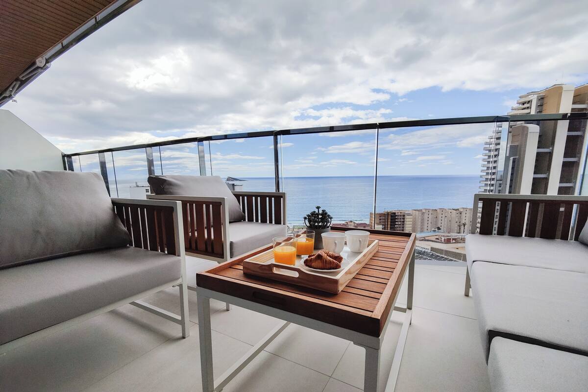 A balcony with a wooden table, two grey sofas, and a tray with croissants, juice, and coffee cups.