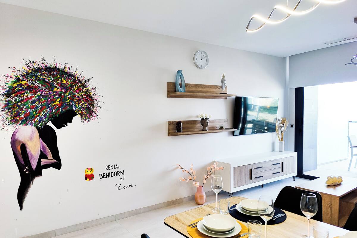 A modern dining area with a wooden table set for two, a TV mounted on the wall, and a decorative wall mural.