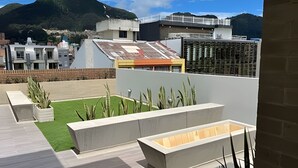 A rooftop terrace with a small lawn, concrete planters, and a view of surrounding buildings and mountains. - Edificio Loft 20-51 (Bogotá)