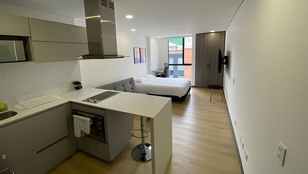 A compact kitchen with a central island, a microwave, and a stove. A bed is visible in the background with a view of buildings outside. - Edificio Loft 20-51 (Bogotá)