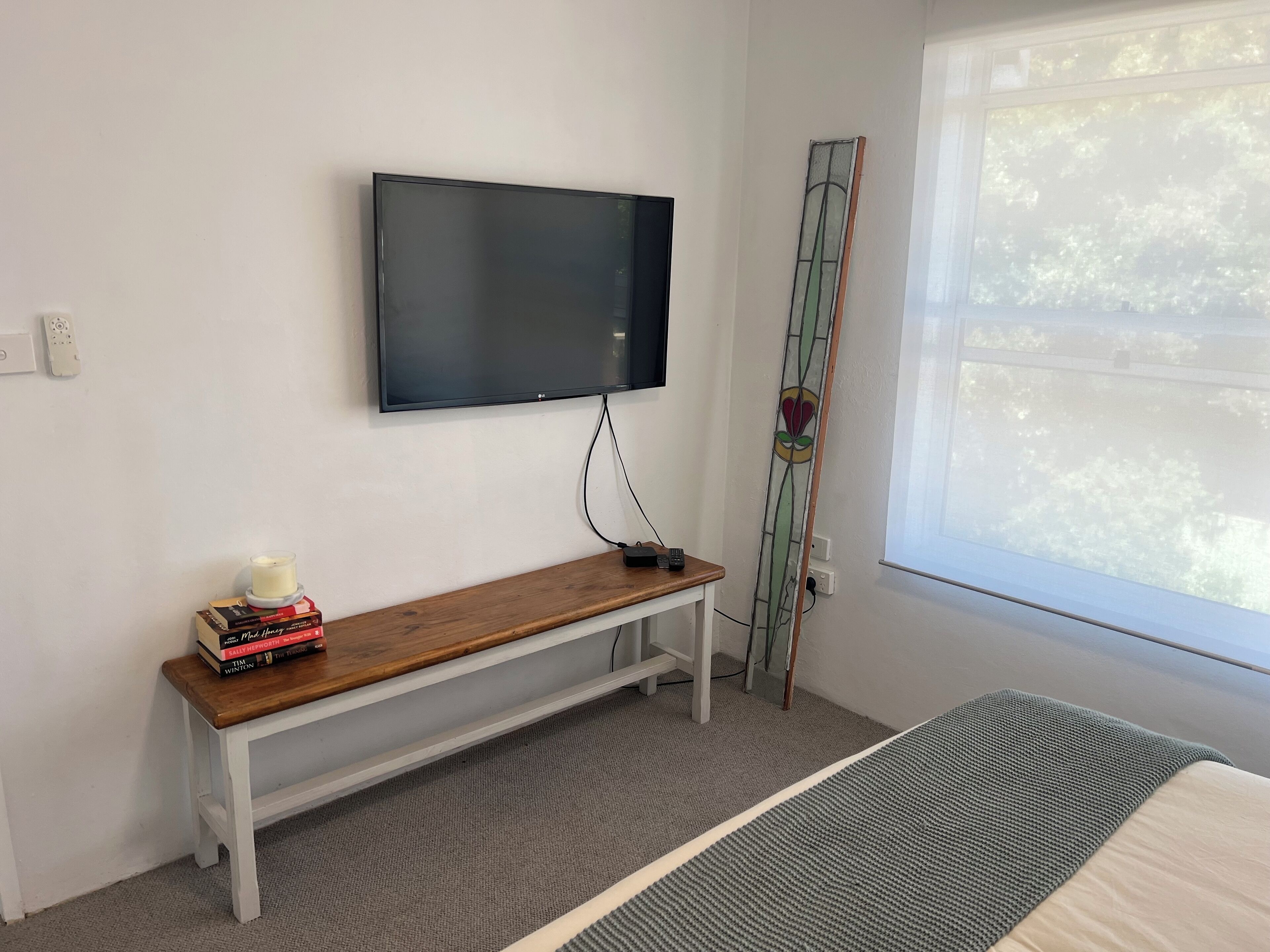 Ett rum med en tv som är monterad på väggen, en träbänk med böcker, en stege och ett fönster med utsikt över träd.