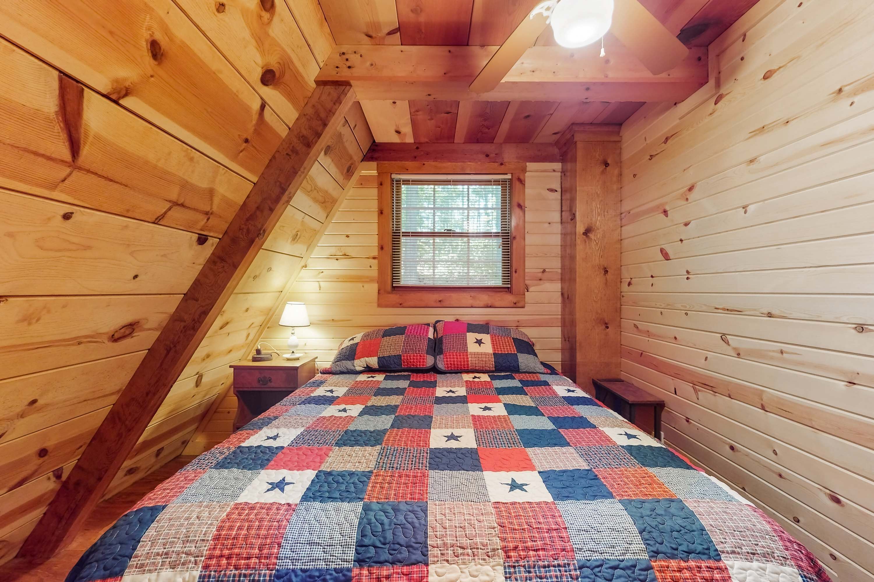 A bedroom with a wooden sloped ceiling, a quilted bedspread, a nightstand, and a window with blinds.