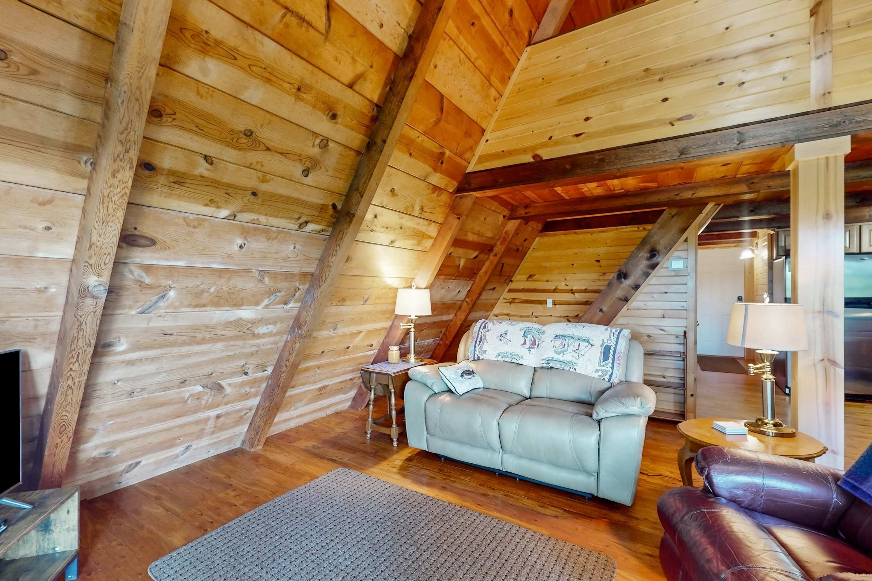 Ein gemütliches Schlafzimmer im Dachgeschoss mit Holzwänden und schrägen Decken. Ein graues Sofa und ein roter Ledersessel stehen an der Wand. Ein kleiner Tisch mit Lampe und Buch ist sichtbar.