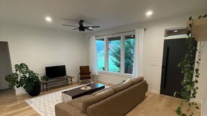Un salon spacieux avec un grand canapé, une table basse et un téléviseur. Il y a une fenêtre avec des rideaux, un ventilateur de plafond et un réfrigérateur.