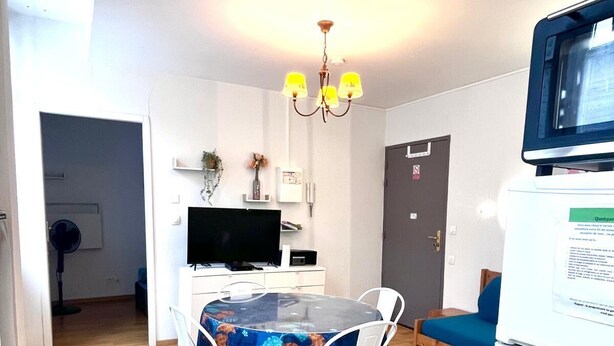 Ein kompakter Wohnraum mit Essbereich, der einen Glastisch und weiße Stühle umfasst, einer Küchenzeile mit Mikrowelle sowie einer kleinen Sitzecke mit blauem Kissen.